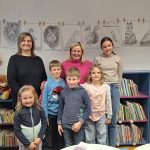 Na fotografiji so udeleženci likovnih delavnic Risanje z Damijano Bijek skupaj z mentorico in organizatorko. Skupina otrok stoji pred knjižnimi policami v knjižnici, za njimi pa so na vrvici razstavljene njihove risbe živali, pritrjene z lesenimi &scaron;čipalkami. Otroci se nasmejani pozirajo ob dveh odraslih, ki sta sodelovali pri izvedbi delavnic. Fotografija ponazarja prijetno ustvarjalno vzdu&scaron;je ter ponos udeležencev ob razstavljenih delih.