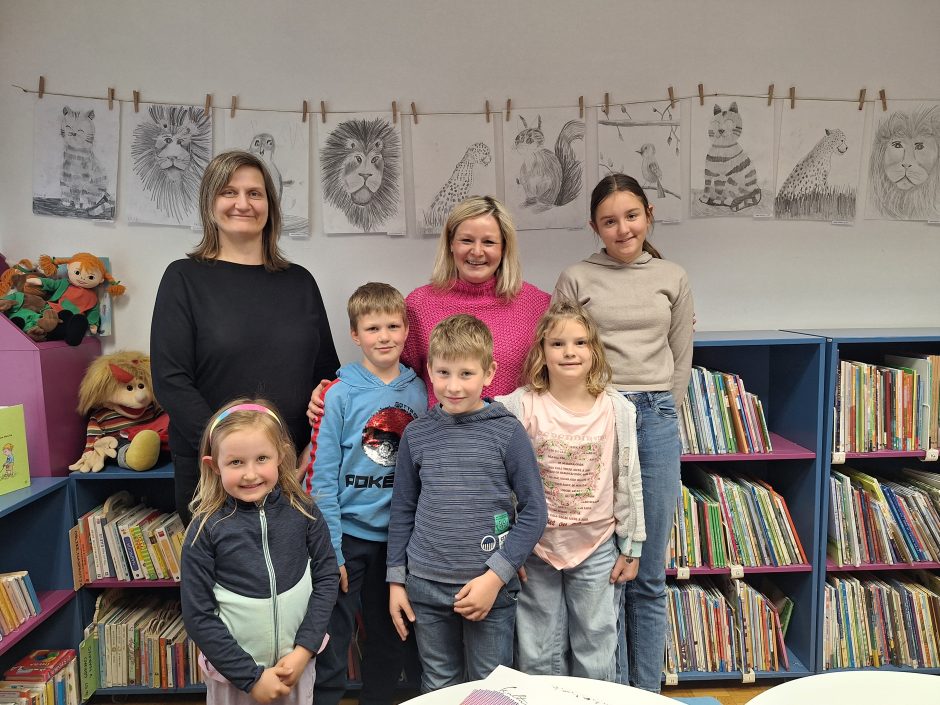 Na fotografiji so udeleženci likovnih delavnic Risanje z Damijano Bijek skupaj z mentorico in organizatorko. Skupina otrok stoji pred knjižnimi policami v knjižnici, za njimi pa so na vrvici razstavljene njihove risbe živali, pritrjene z lesenimi &scaron;čipalkami. Otroci se nasmejani pozirajo ob dveh odraslih, ki sta sodelovali pri izvedbi delavnic. Fotografija ponazarja prijetno ustvarjalno vzdu&scaron;je ter ponos udeležencev ob razstavljenih delih.