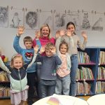 Na fotografiji so udeleženci likovnih delavnic Risanje z Damijano Bijek skupaj z mentorico. Skupina otrok stoji pred knjižnimi policami v knjižnici, za njimi pa so na vrvici razstavljene njihove risbe živali, pritrjene z lesenimi &scaron;čipalkami. Otroci se nasmejani pozirajo ob dveh odraslih, ki sta sodelovali pri izvedbi delavnic. Fotografija ponazarja prijetno ustvarjalno vzdu&scaron;je ter ponos udeležencev ob razstavljenih delih.