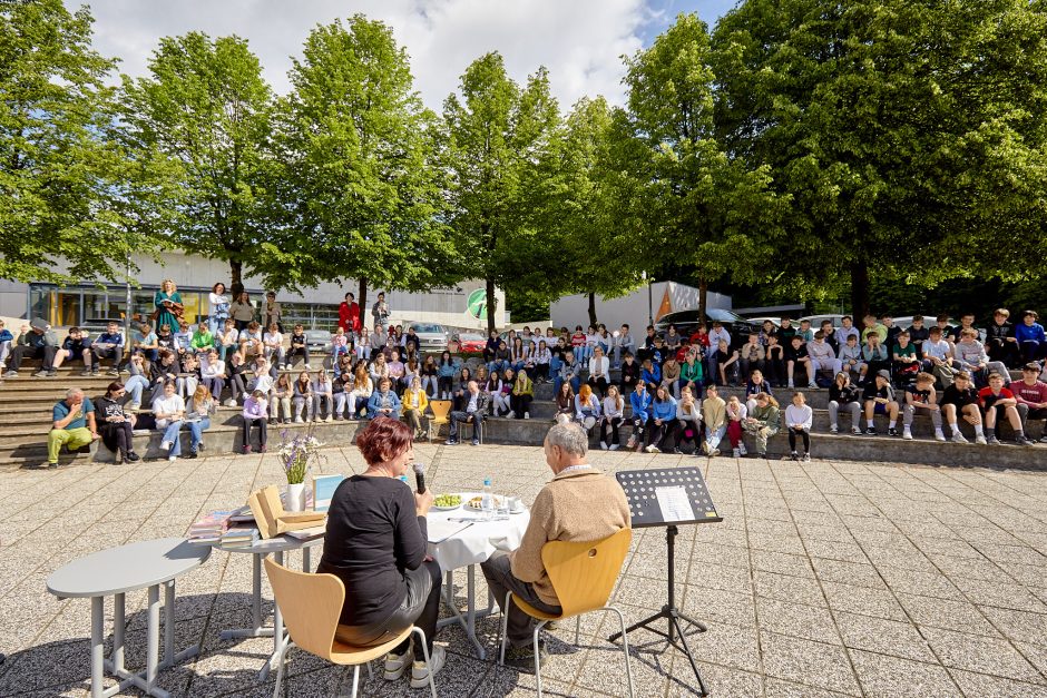 Na fotografiji dva govorca sedita za mizo na prostem in nagovarjata veliko skupino mladih, zbranih na stopničastem amfiteatru pod zelenimi drevesi v sončnem vremenu.