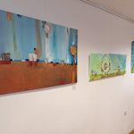 Na beli galerijski steni visijo tri abstraktne slike v živih modrih, zelenih in toplih zemeljskih tonih. Dela prikazujejo stilizirane pokrajine in krožne motive ter ustvarjajo umirjeno, sodobno razstavno postavitev.